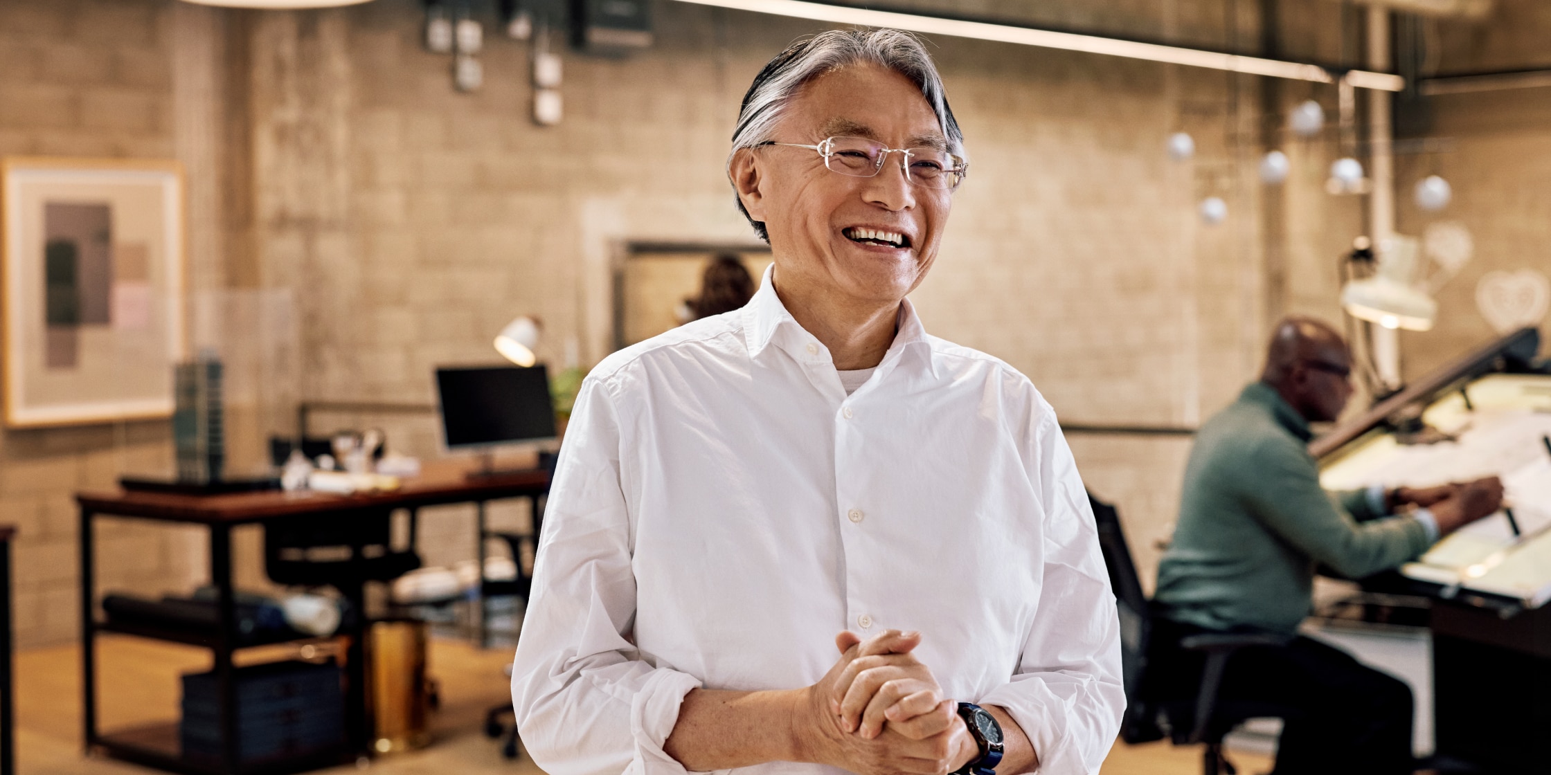 A man in a white button-down shirt stands in the foreground of an office setting.