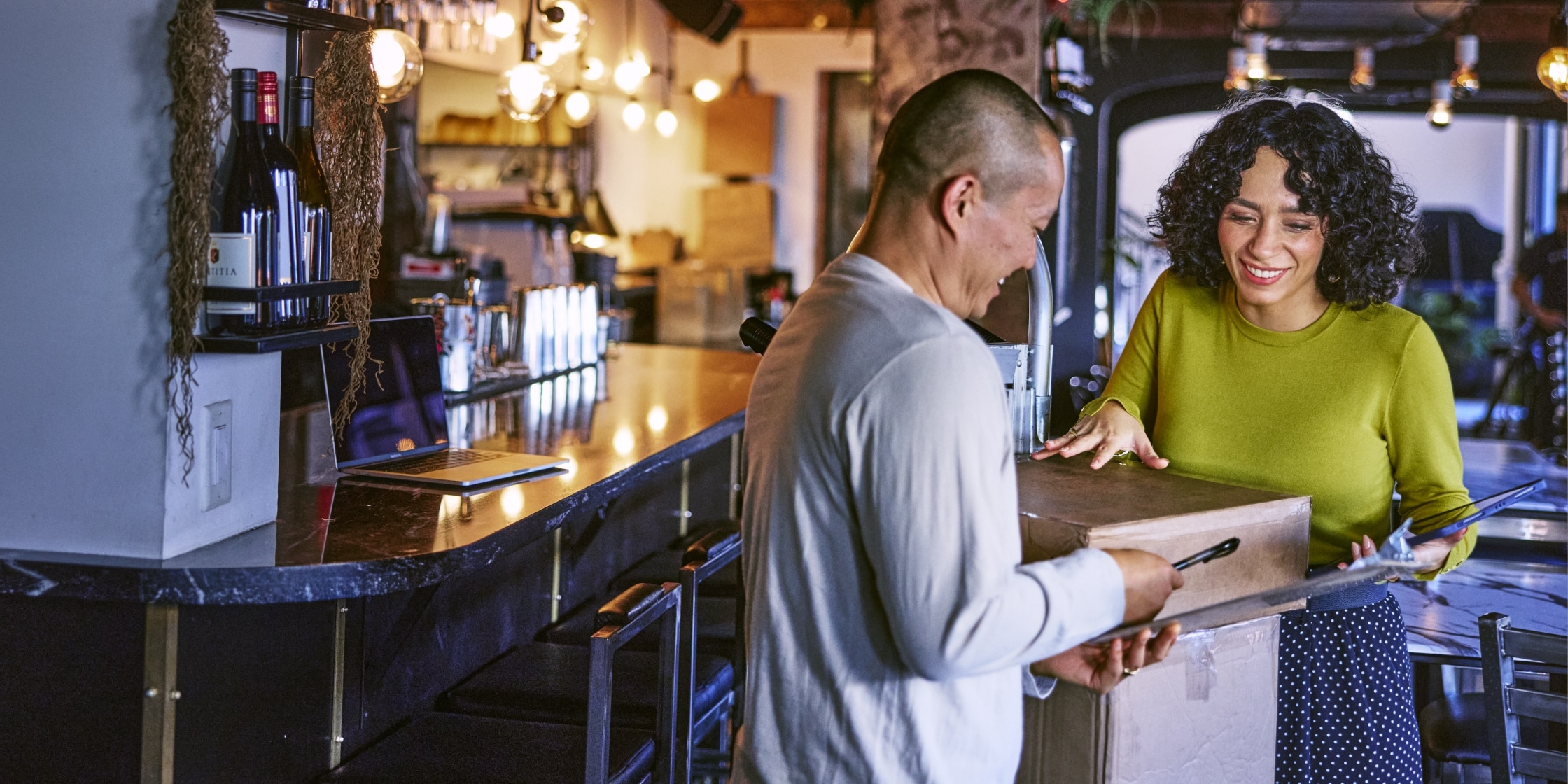 A delivery man passes a pen to a woman so she can sign a receiving form. The woman is wearing a green sweater and a blue polka-dot skirt. There are boxes between them.