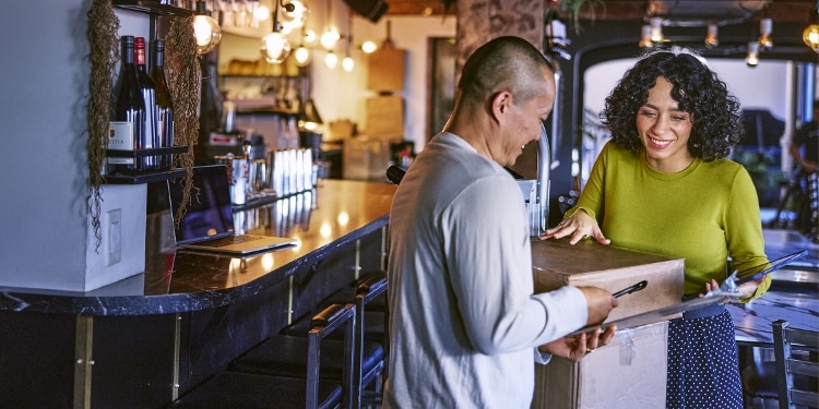 A delivery man passes a pen to a woman so she can sign a receiving form. The woman is wearing a green sweater and a blue polka-dot skirt. There are boxes between them.
