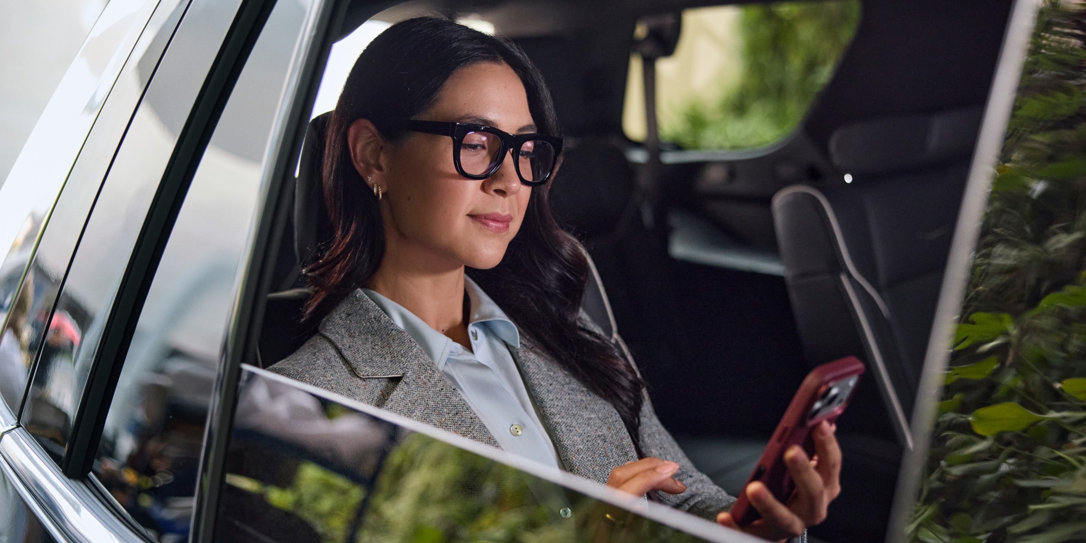 A woman with thick black glasses is sitting in the backseat of a car and looking at her red mobile phone.