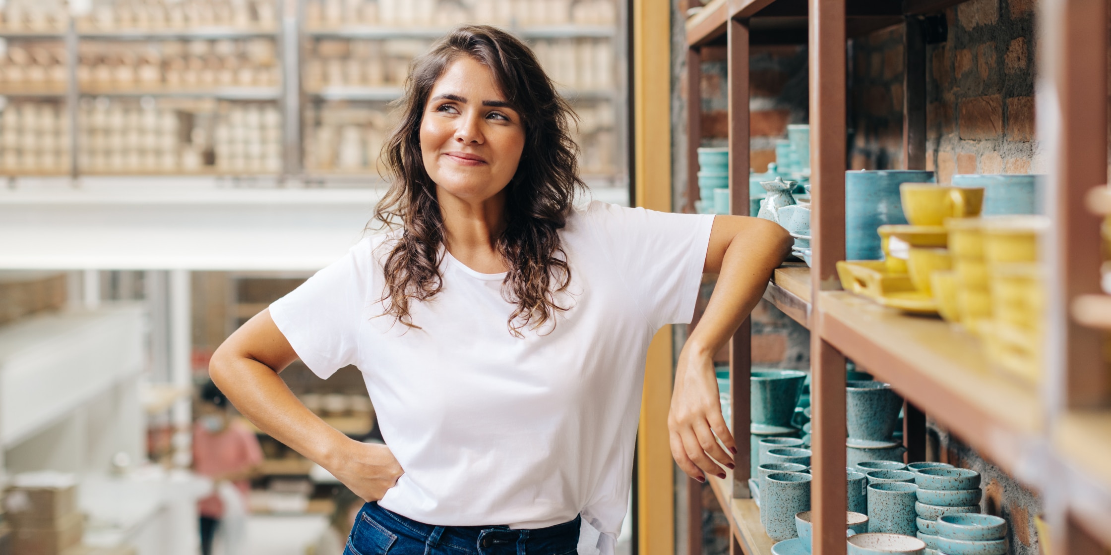 A woman in a white T-shirt smiles while leaning against shelves full of pottery.