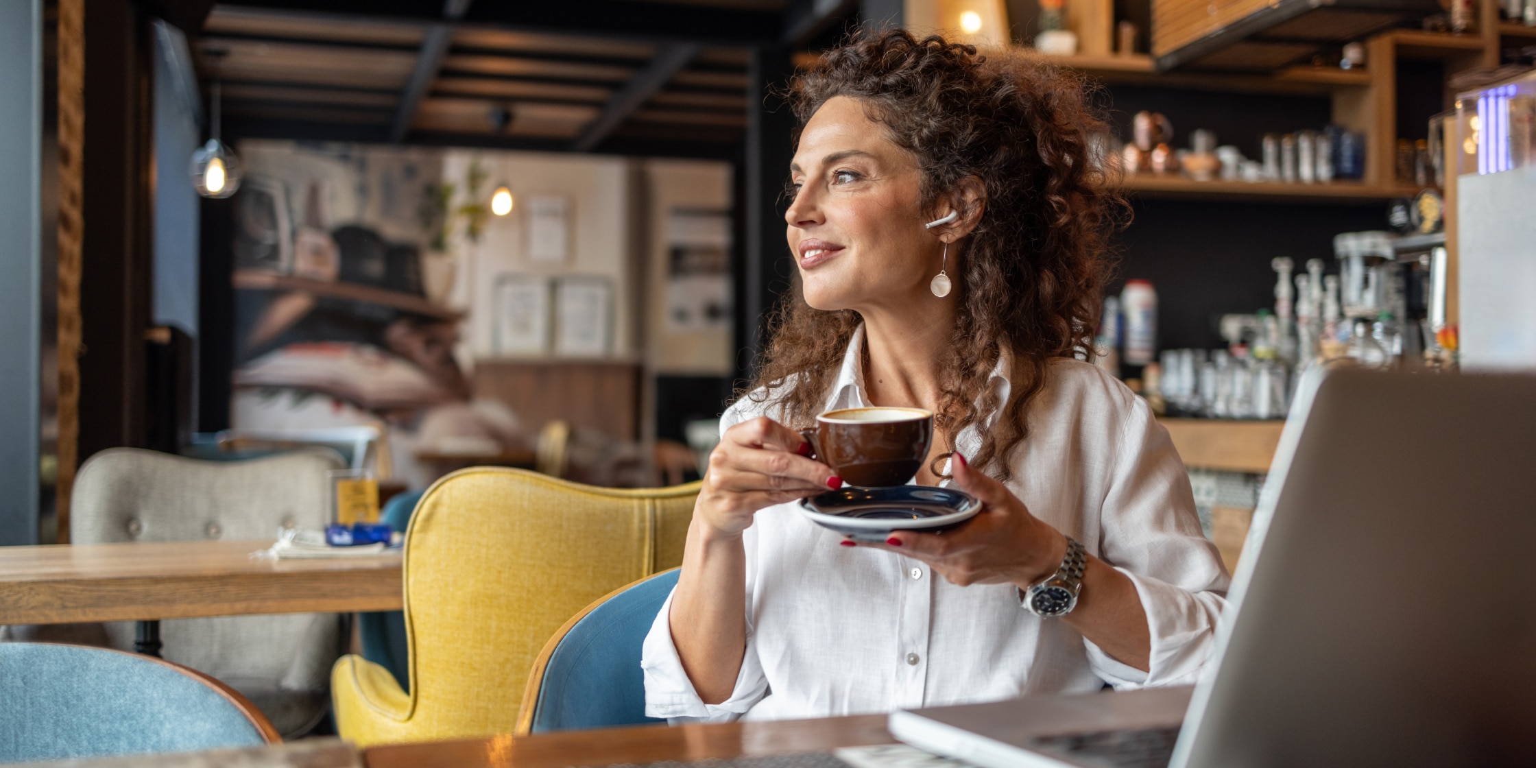 A woman sits at a table in a cafe. She has a latte in her hands and a laptop is open in front of her.