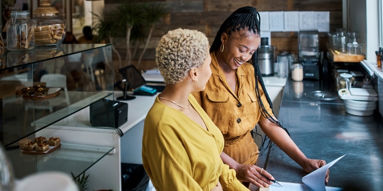 Two women stand behind the counter of a cafe looking over a spreadsheet.