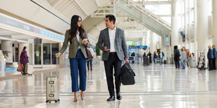 Two individuals who appear to be traveling for business walking through an airport terminal with their carry-on luggage while chatting.