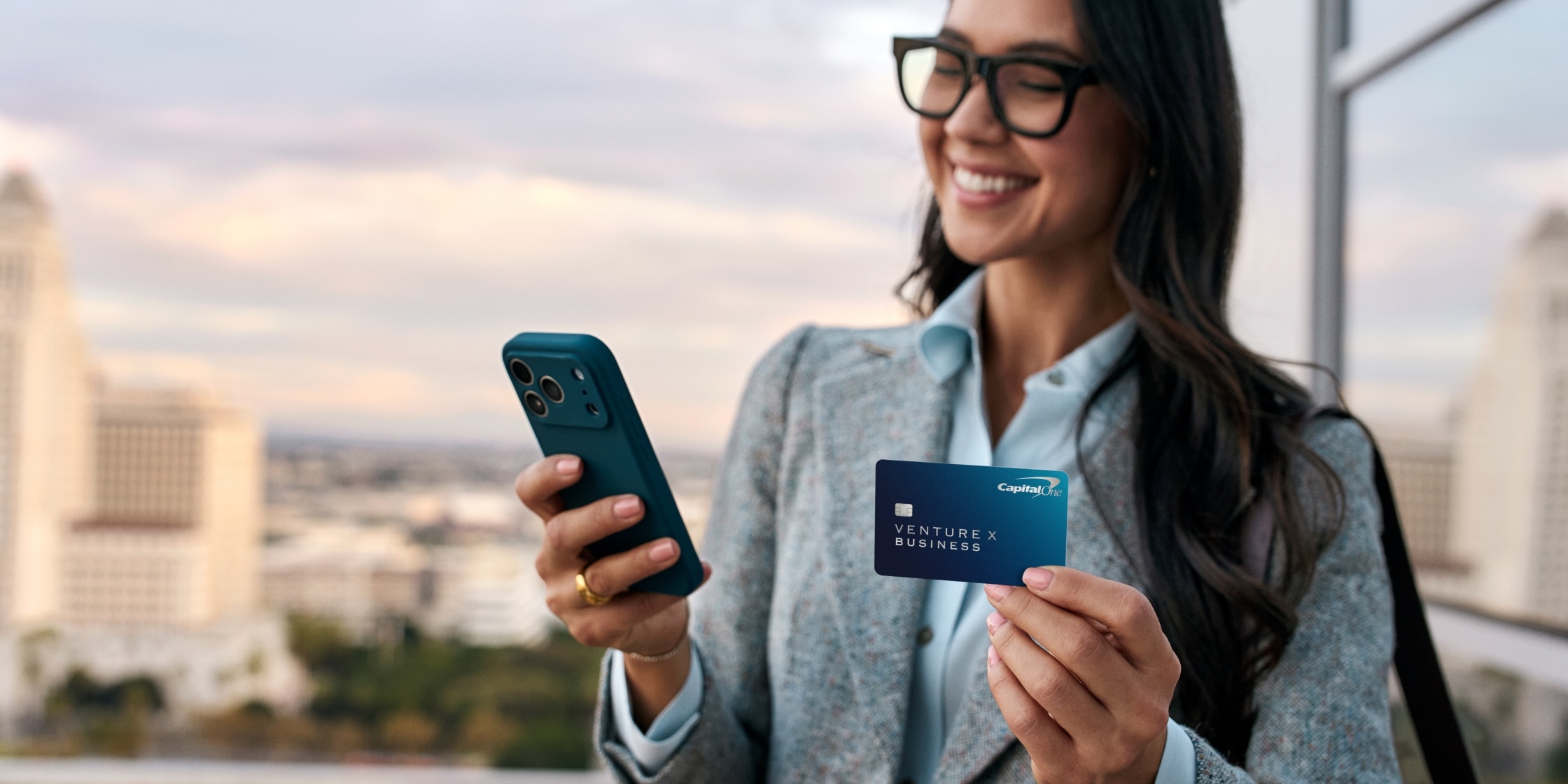 A woman in a blazer holds a blue Venture X Business card in one hand and her mobile phone in the other. There’s a city view behind her.