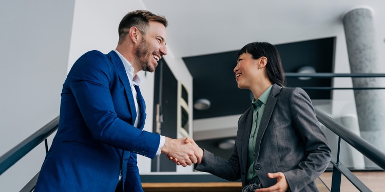 A man in a blue business suit and a woman in a green shirt with a charcoal blazer shake hands while standing in an office stairwell.