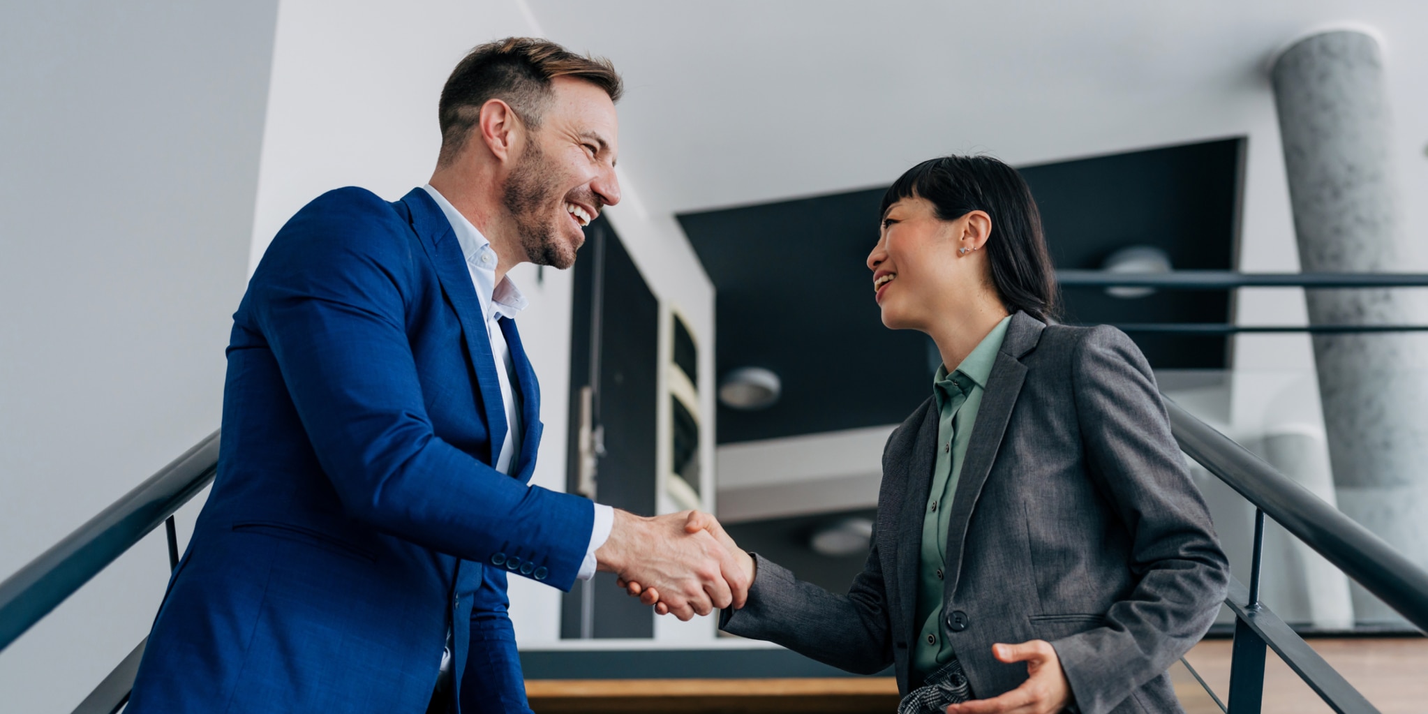 A man in a blue business suit and a woman in a green shirt with a charcoal blazer shake hands while standing in an office stairwell.