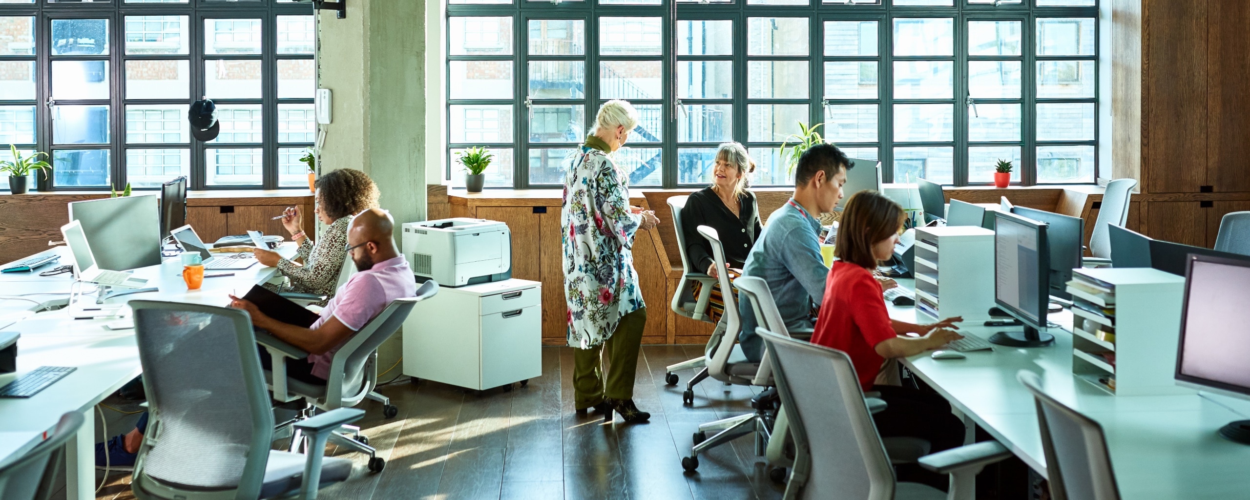 A team of employees in an open plan office sit at their desks and have conversations.