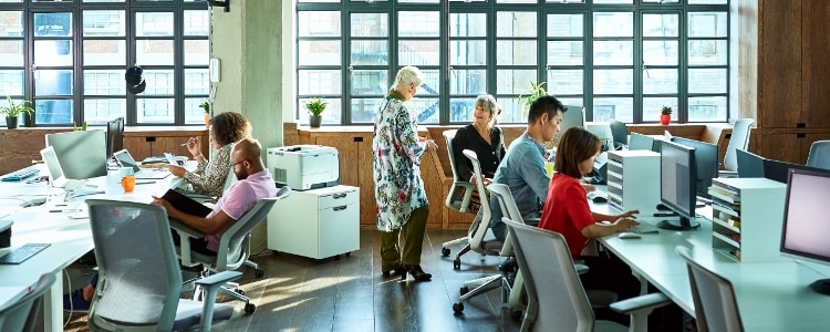 A team of employees in an open plan office sit at their desks and have conversations.