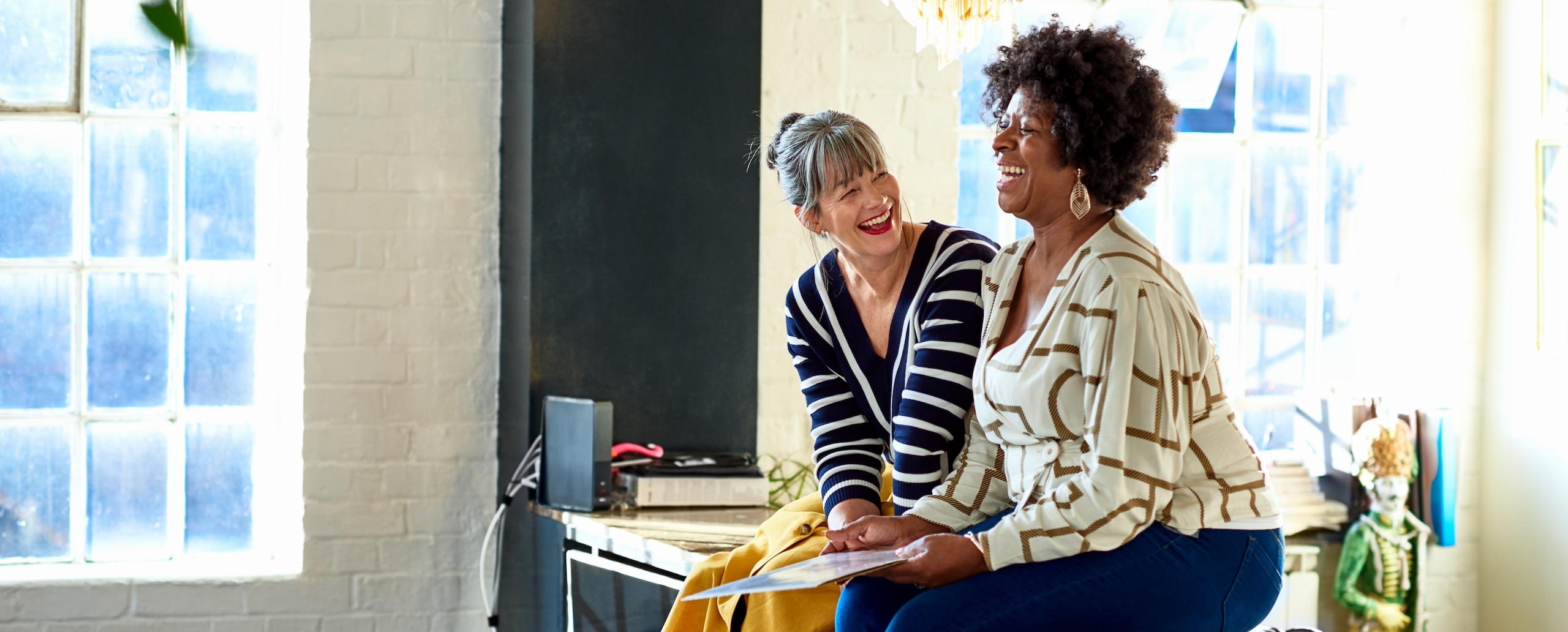 Two women sitting next to each other at a desk while laughing and working on a project together.
