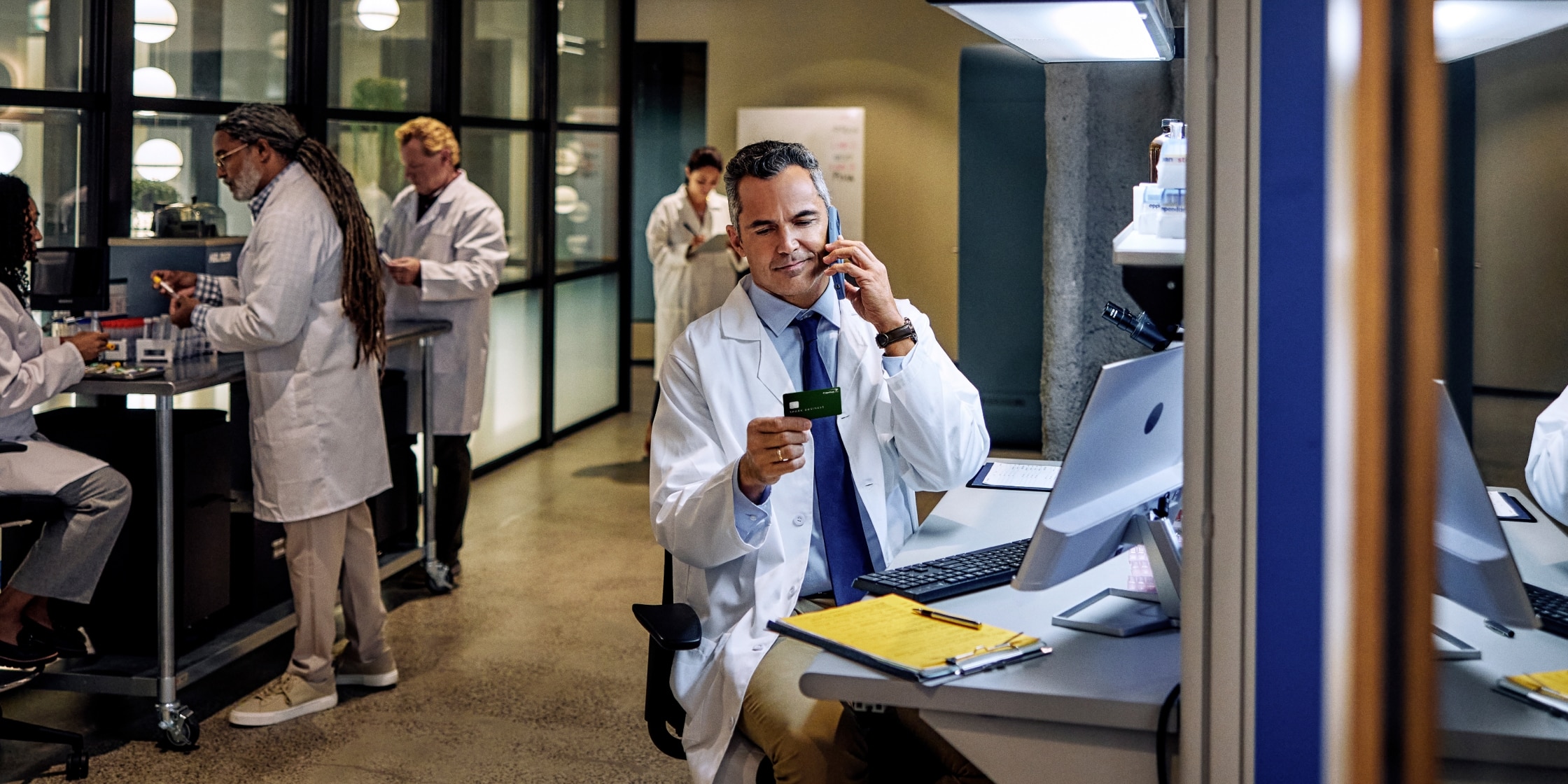An image of a laboratory. There are people in white lab coats at workstations. One of them is sitting down while on the phone. He’s holding a Capital One business card in his other hand.