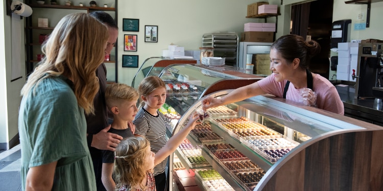 A shop employee hands a treat across a glass display case to a young child while a parent and two other children watch inside a small dessert shop.