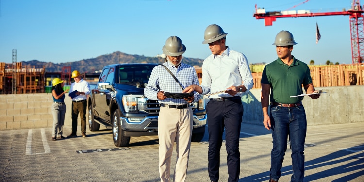 Three men stand together in a construction site. They’re all wearing hardhats and holding clipboards. There’s a truck in the background with others standing beside it.