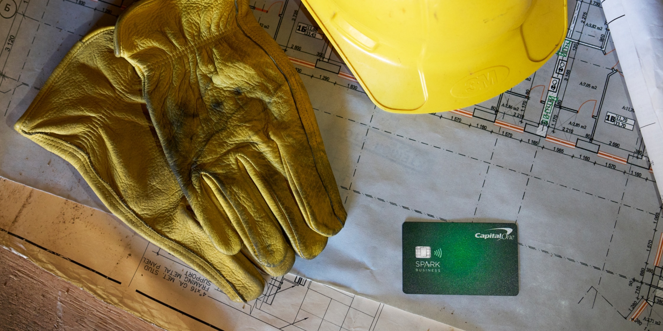 An overhead view of a construction work station. There are blueprints on the table, a yellow hard hat, yellow work gloves and a green Capital One Spark business card.