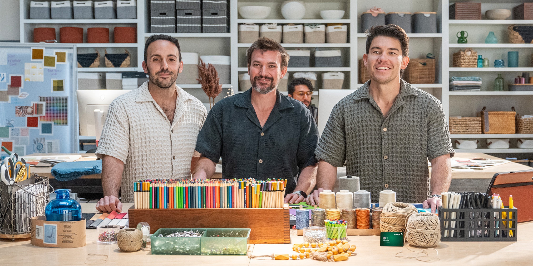 Brandon, Bruno and Steven, the founders of Sand Cloud, stand in front of a worktable covered with threads and colored pencils.