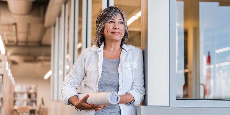 A woman in a gray shirt stands in a hallway holding papers.