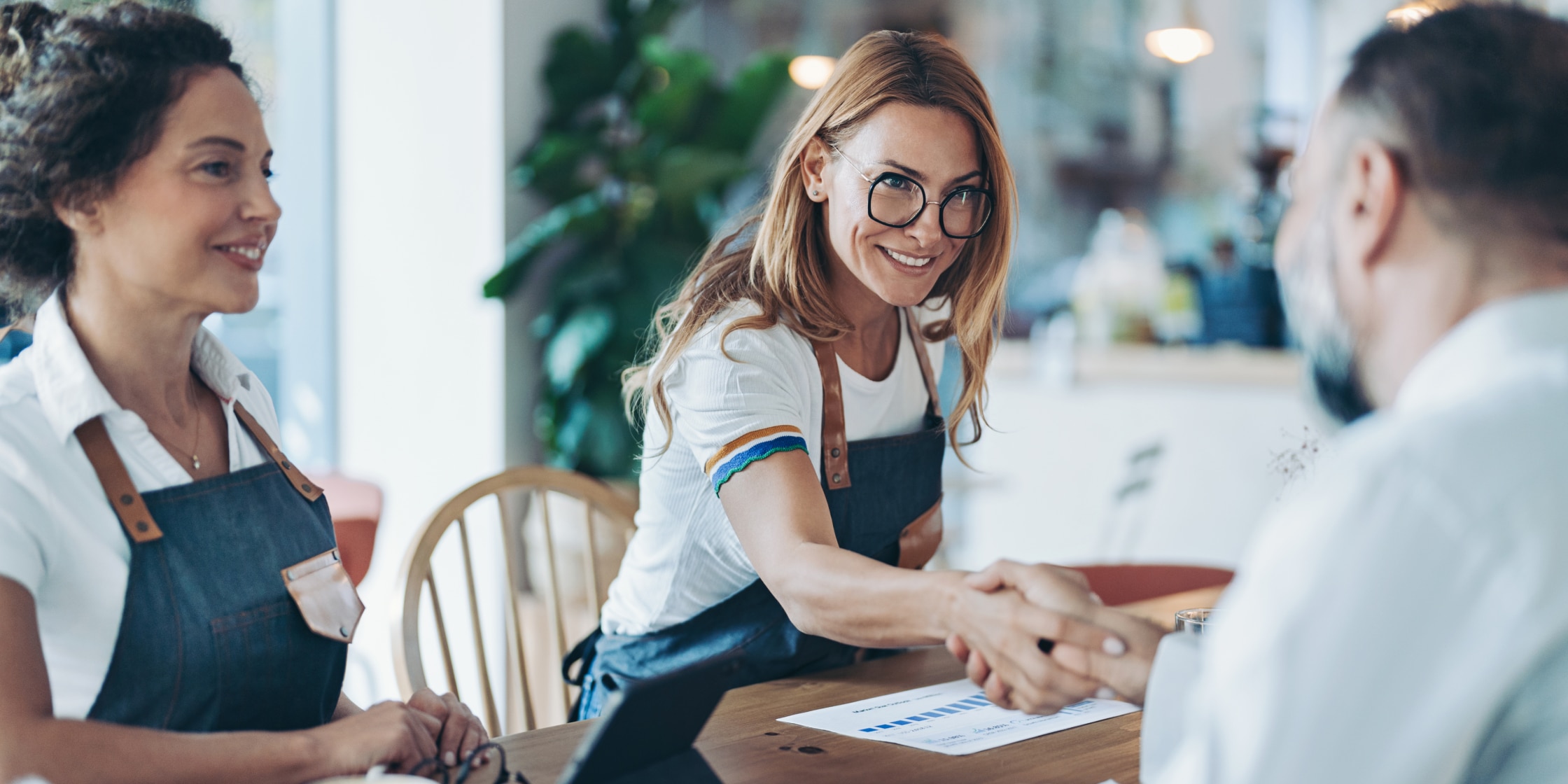 Two women in aprons sit opposite of a man at a table in a restaurant. One woman is shaking the man’s hand, and there’s paperwork on the table between them.