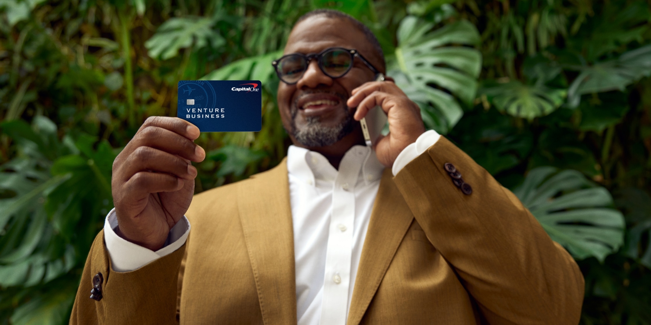 A man in a tan blazer is holding his phone against his ear in one hand and a blue Capital One Venture X Business card in the other. There are palm trees behind him.