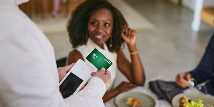 A woman sits at a restaurant table in the background, while a waiter taps her green Capital One Spark Cash Plus card against a credit card reader in the foreground.