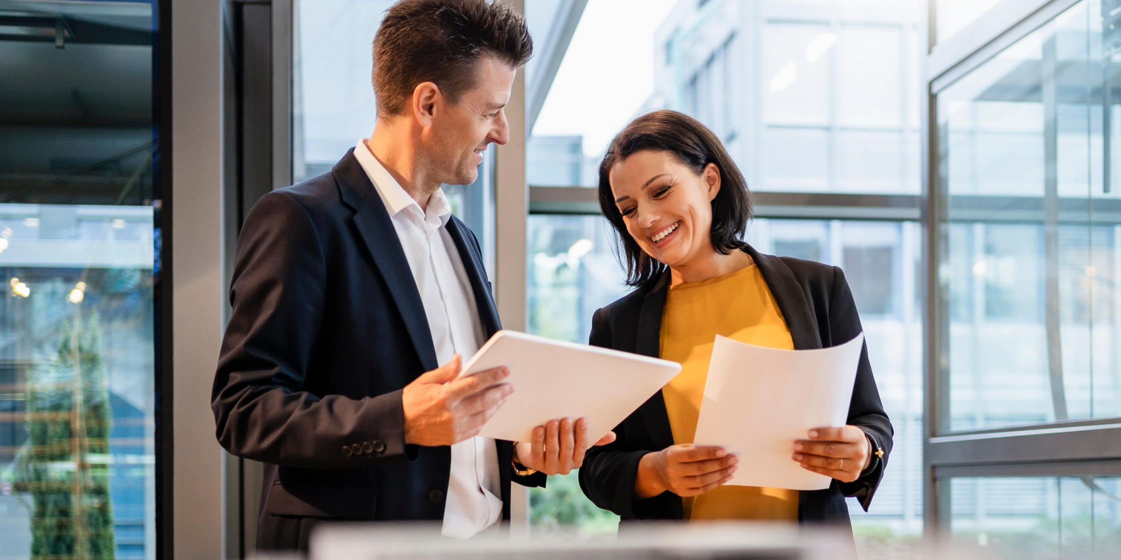 A woman and a man stand beside each other in an office. Both are holding papers and showing them to each other.