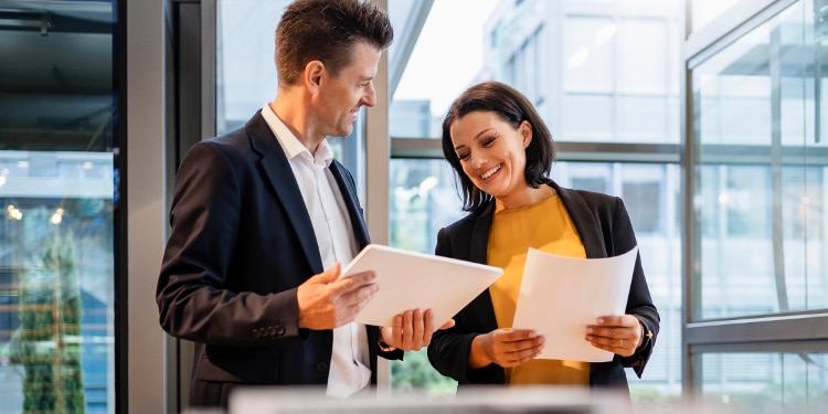 A woman and a man stand beside each other in an office. Both are holding papers and showing them to each other.