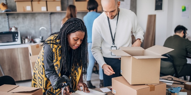 A woman and a man work at an office table together. The woman is wearing a yellow sweater vest and is looking in a notebook, while the man looks over her shoulder and opens a cardboard box. Other people are in the background working as well.