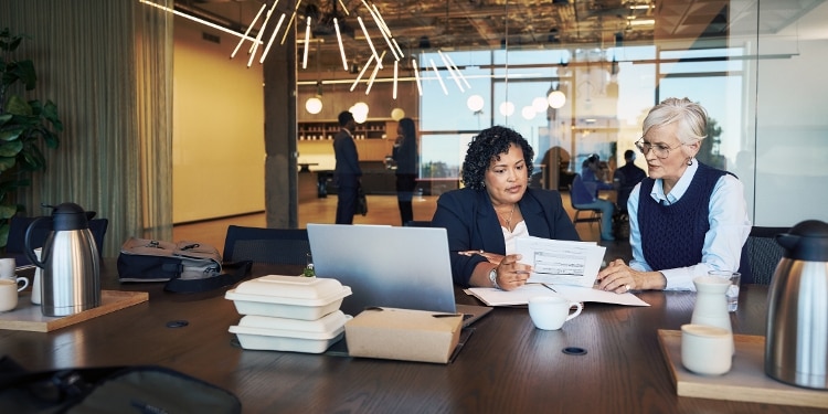 Two women sit at an office conference table, looking over documents together.
