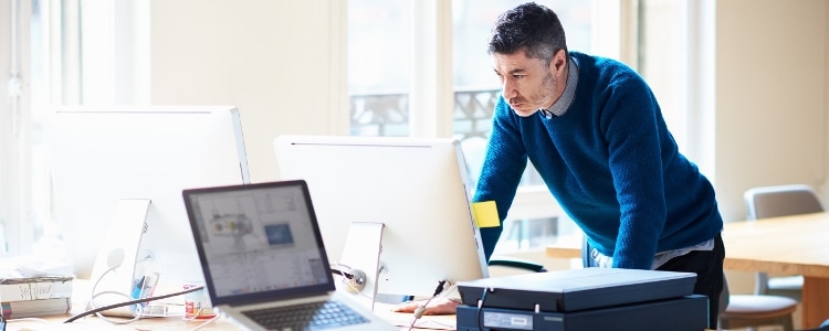 Man in an office space leaning against a desk looking at a computer monitor