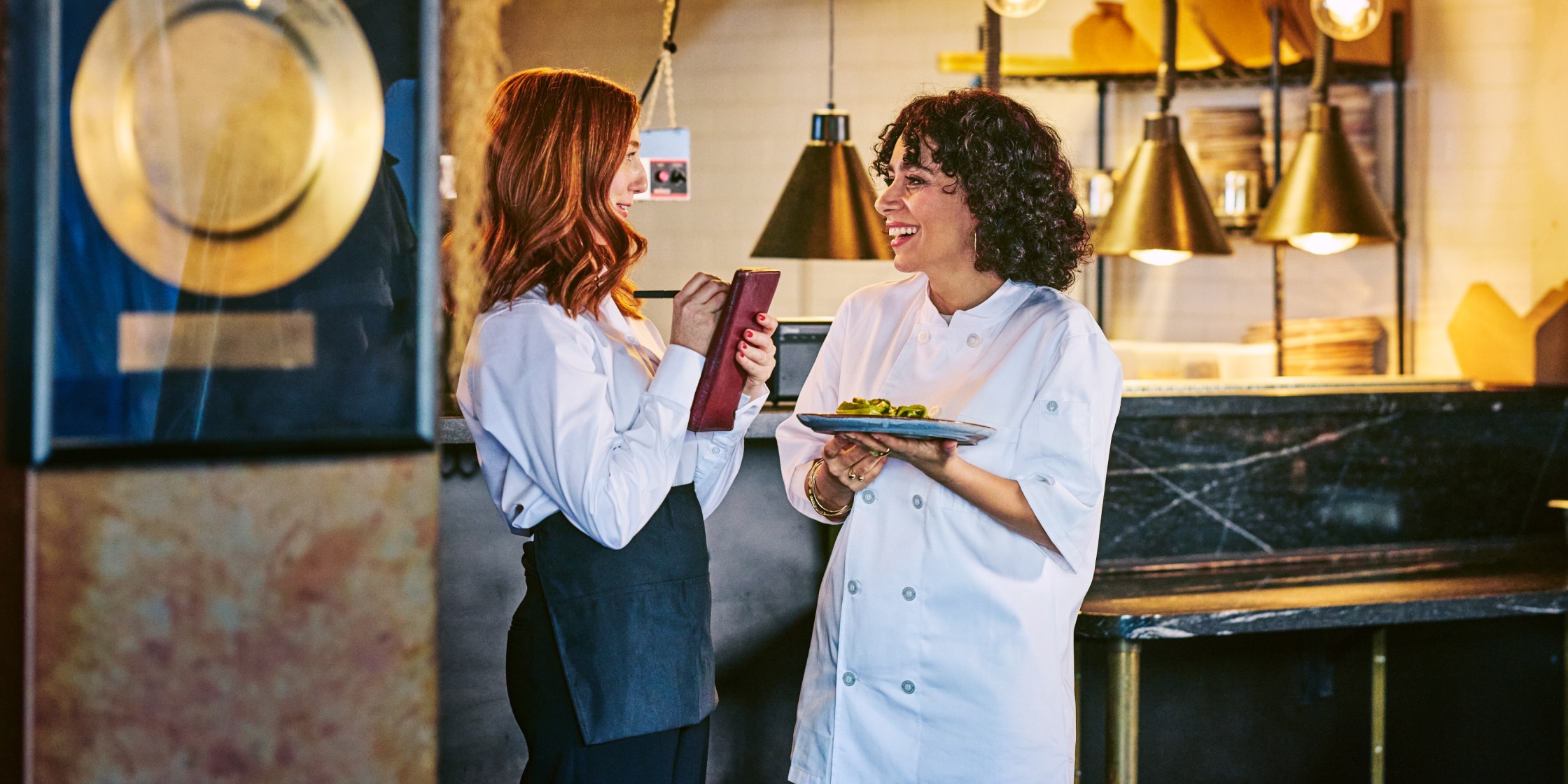A woman in a chef’s coat stands in a restaurant. She’s holding a plate of food and talking to a server writing something down on a pad.