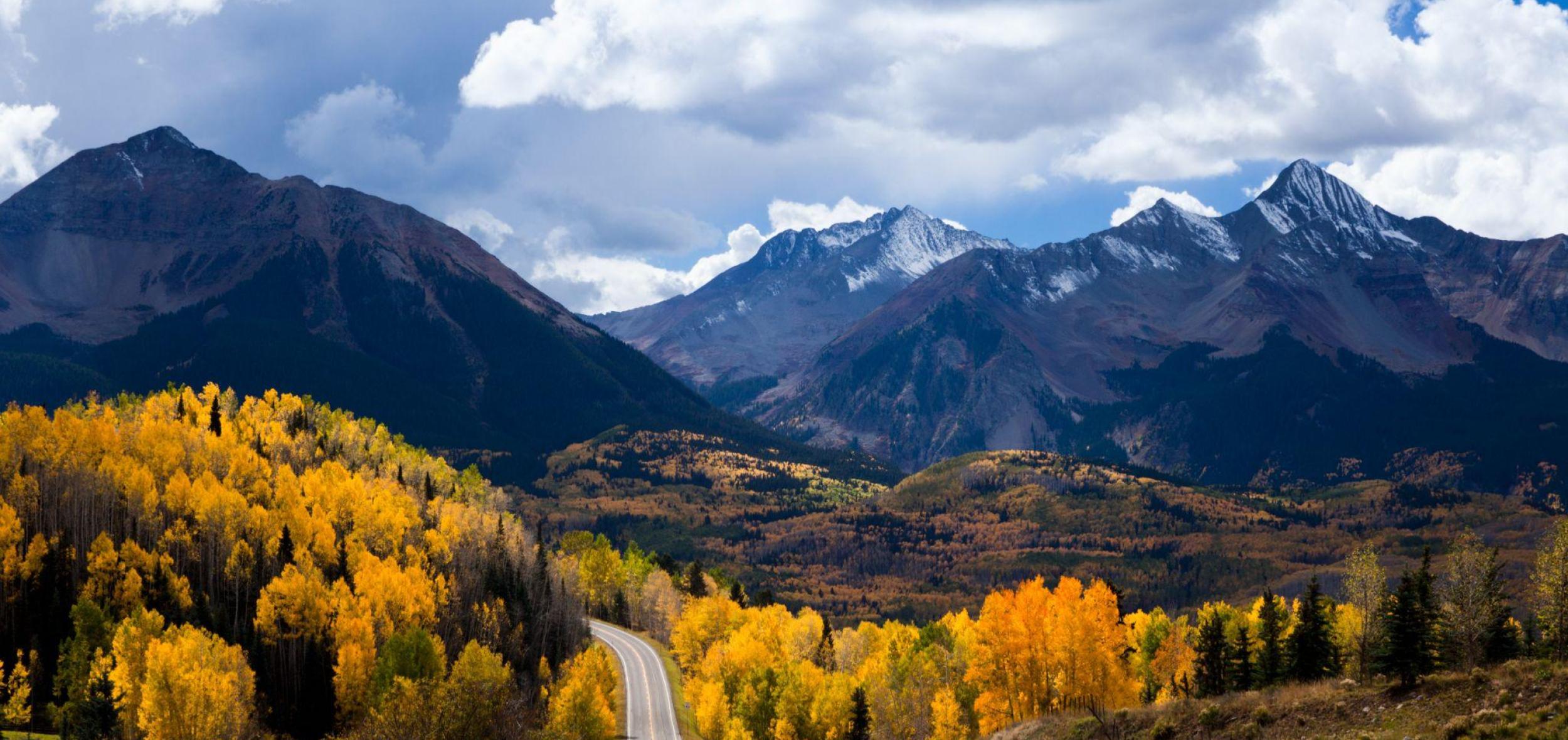 A wide panoramic shot of a mountain range under a cloudy sky, featuring brilliant golden autumn foliage covering the foothills in the foreground.