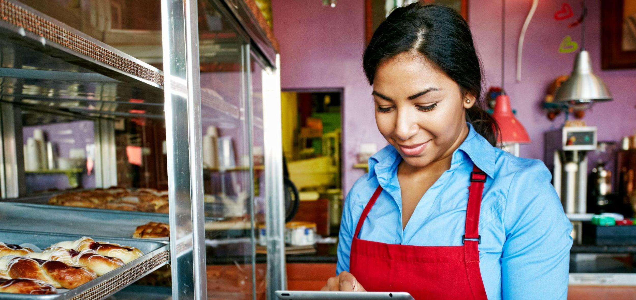 A smiling female small business owner wearing a red apron and blue shirt uses a digital tablet inside her bakery, with trays of fresh pastries visible in a display case.