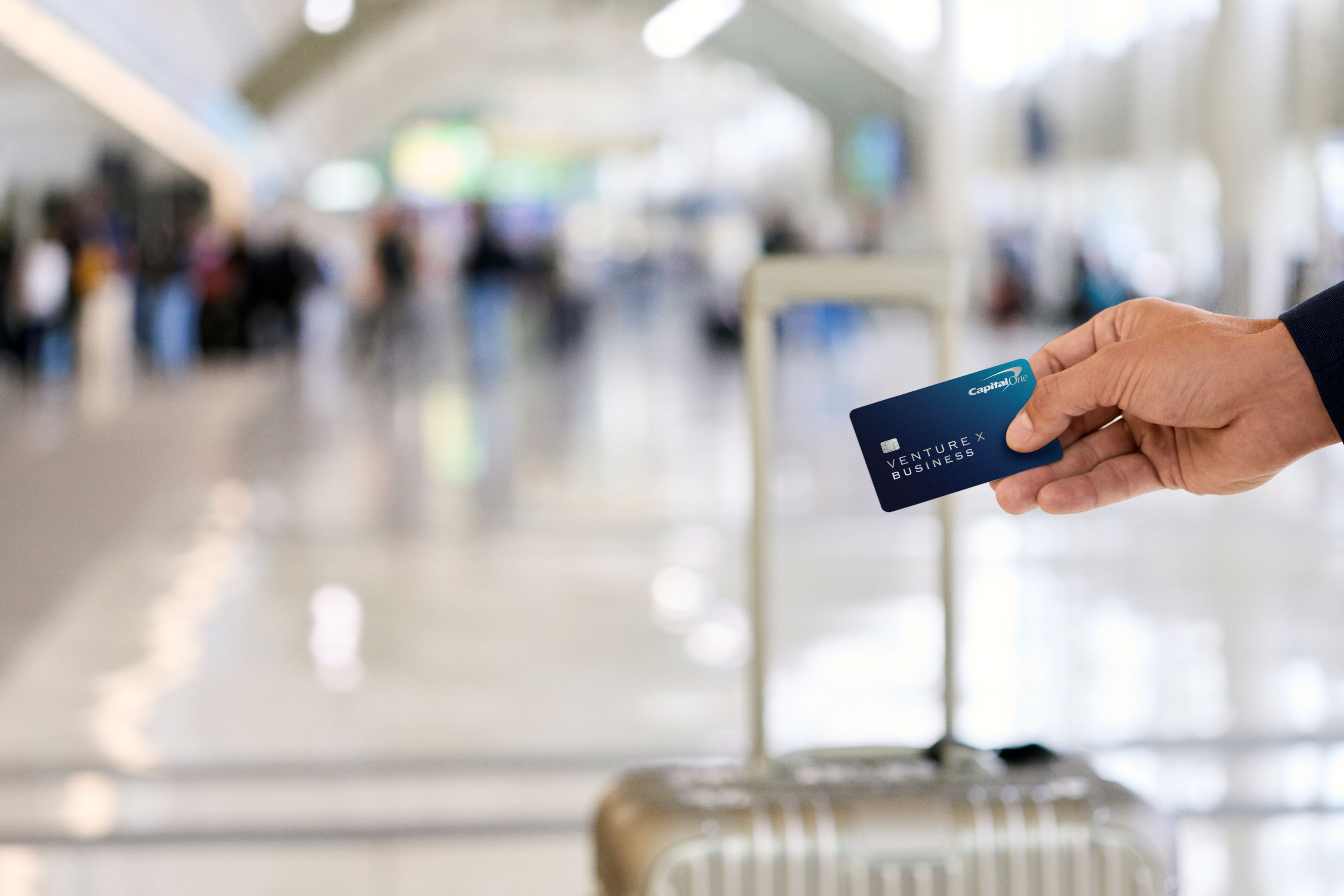 A person's hand holding a dark blue Capital One Venture X Business credit card in front of a silver luggage handle, with a blurred, brightly lit airport terminal in the background.
