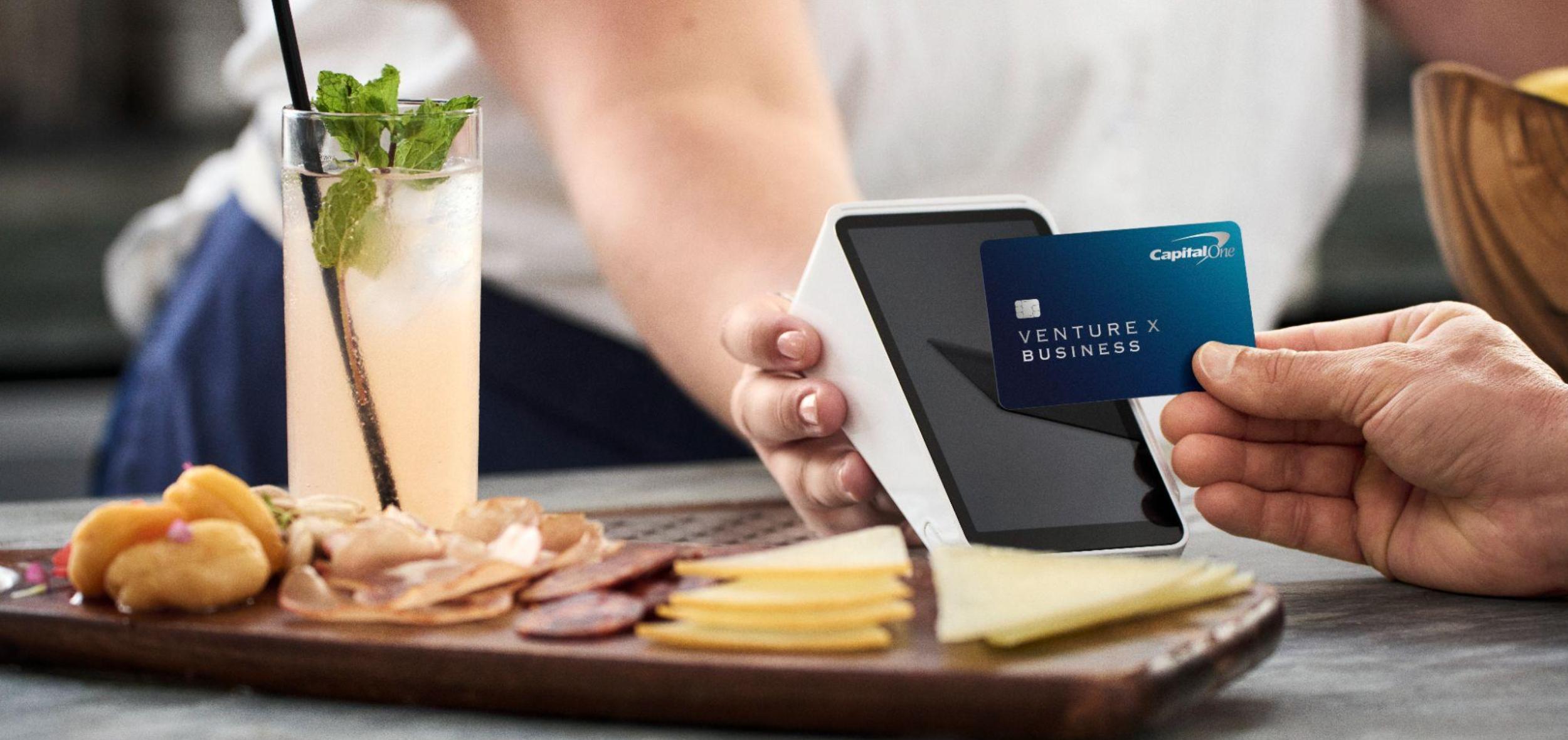 A customer taps a blue Capital One Venture X Business card onto a handheld contactless terminal at a restaurant table with a cocktail and a charcuterie board.