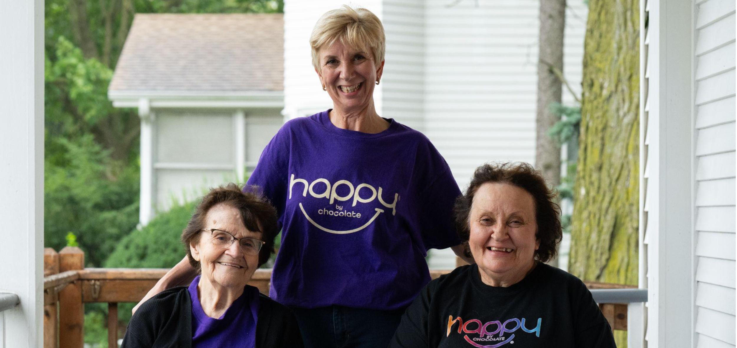 Marcy Vieau-Goetz of Happy by Chocolate stands smiling on an outdoor porch between two seated women wearing branded company shirts.