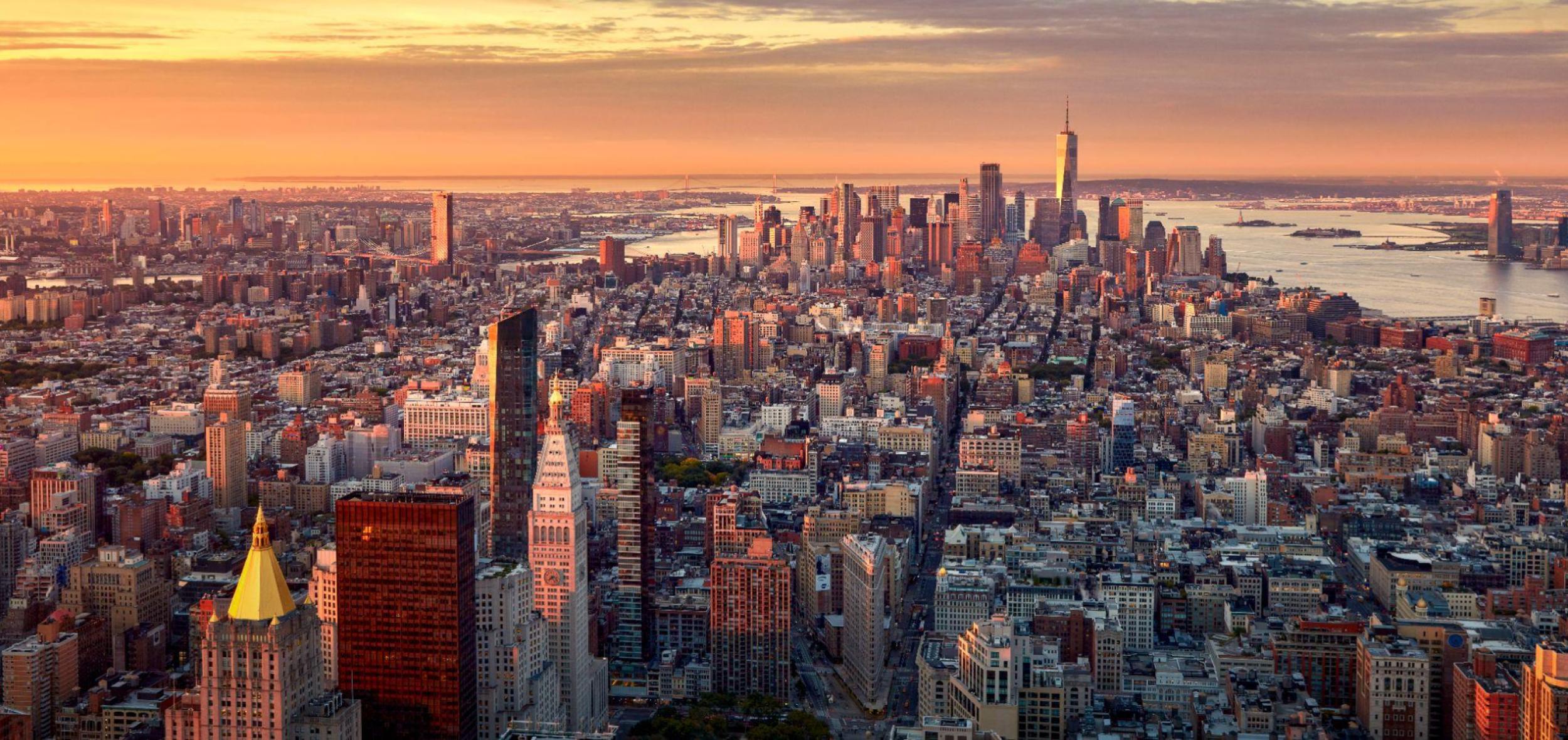 High-angle aerial view of the New York City skyline at sunset, with golden light reflecting off skyscrapers stretching south toward One World Trade Center.