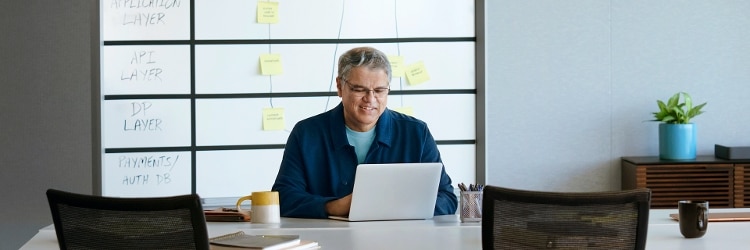 Man in navy shirt sitting in front of whiteboard with a computer on his desk