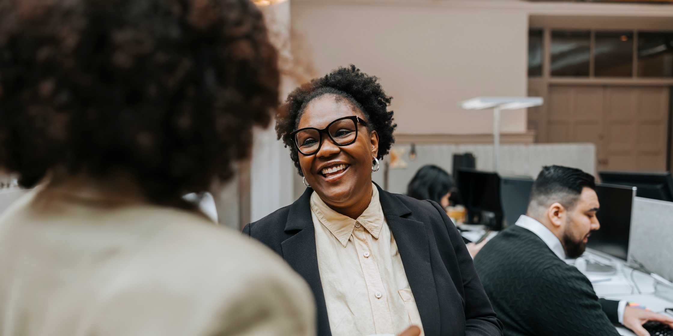 Woman smiles as she converses with another woman, people work at desks in background