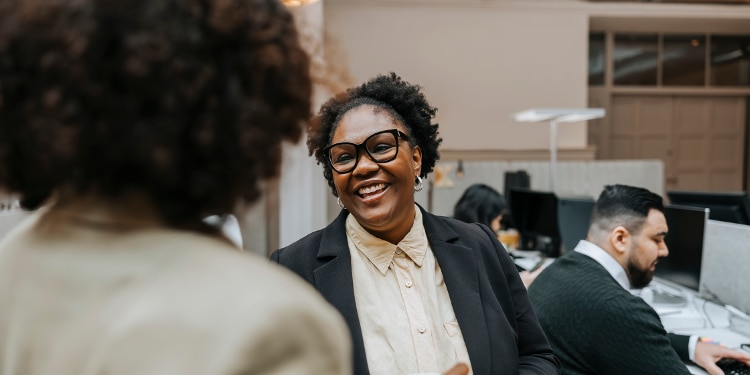 Woman smiles as she converses with another woman, people work at desks in background