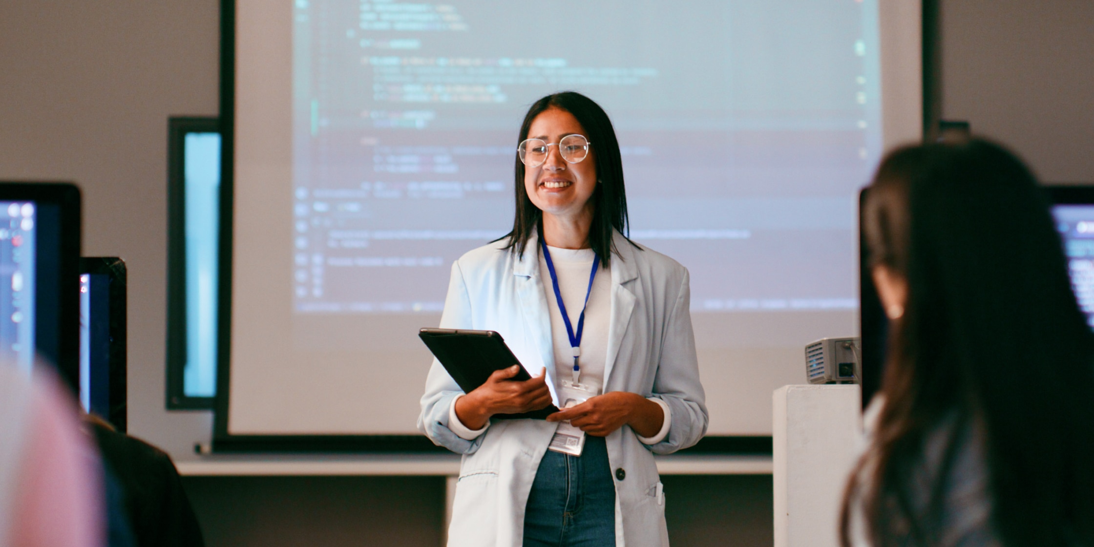 Smiling woman holding tablet stands in front of projector with lines of code on the screen as students look on
