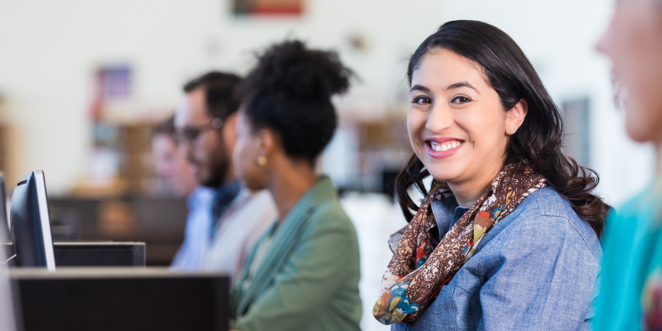 female with dark hair and a brown scarf sitting at a desk smiling towards the camera with other people blurred in the background
