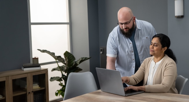 Woman sitting at desk and man standing behind her look at laptop screen