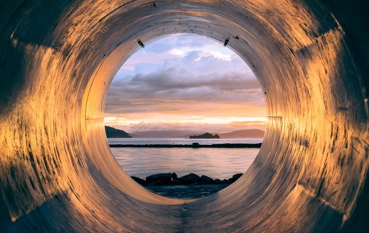 large silver tube with blue ocean, dark mountains, and cloudy sky in distance