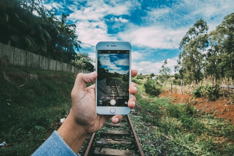 hand holding iphone with picture of railroad and green grass on it, with blue sky and green grass and tree scenary behind it