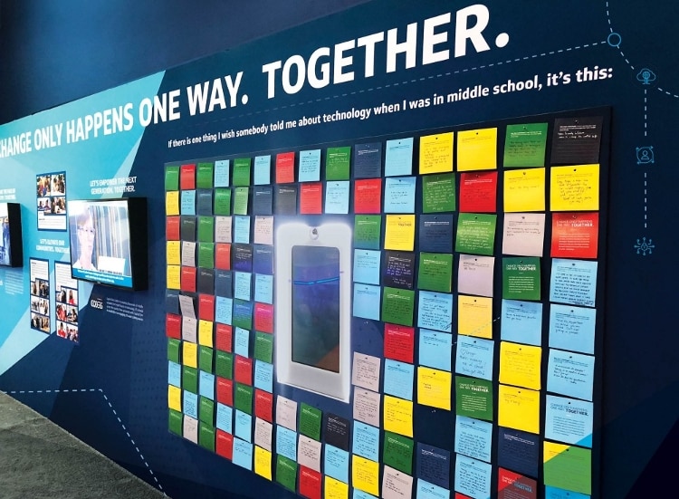 red, blue, pink, yellow, and green sticky notes with black writing taped to conference booth wall with illuminated screens and white block letters