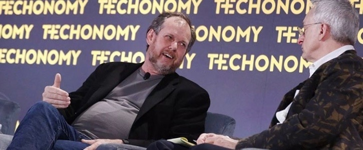 man in black sports jacket and grey shirt sitting down and in conversation with man with grey hair and glasses wearing black sports jacket and white collared shirt. there is a purple wall with yellow block letters behind them