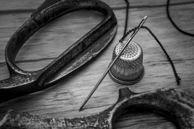 greyscale image of sewing needle with black thread resting on a steel thimble, next to the handles of a black pair of scissors