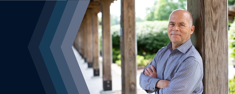 Bernard Golden wearing grey shirt with white lines, leaning against a wooden pillar. more wooden pillars are behind him. there are grey and navy cocentric arrows on the left side of the image, pointing from right to left.