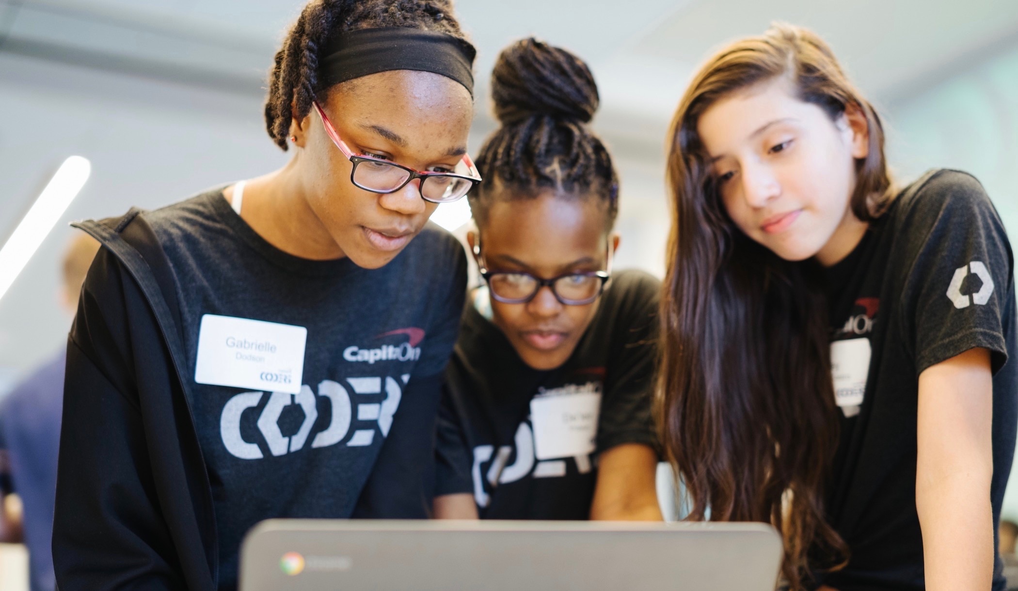 3 young girls wearing black coders shirts with white nametags, working together in front of an open silver laptop