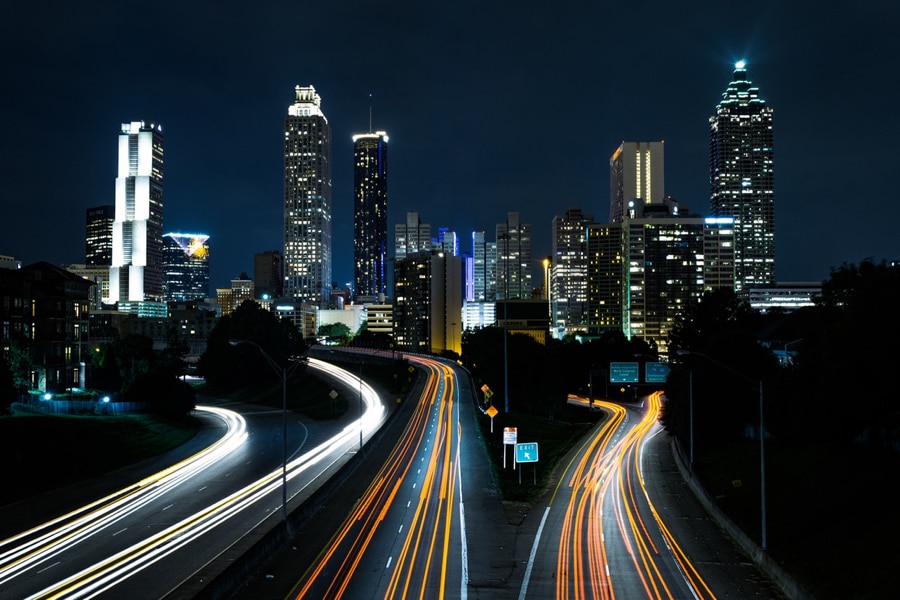night photo of 3 highway lanes with white and orange lines of light going towards a city with skyscrapers with white lights shining through windows
