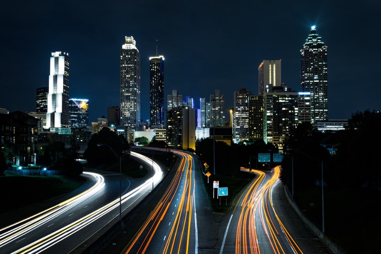night photo of 3 highway lanes with white and orange lines of light going towards a city with skyscrapers with white lights shining through windows